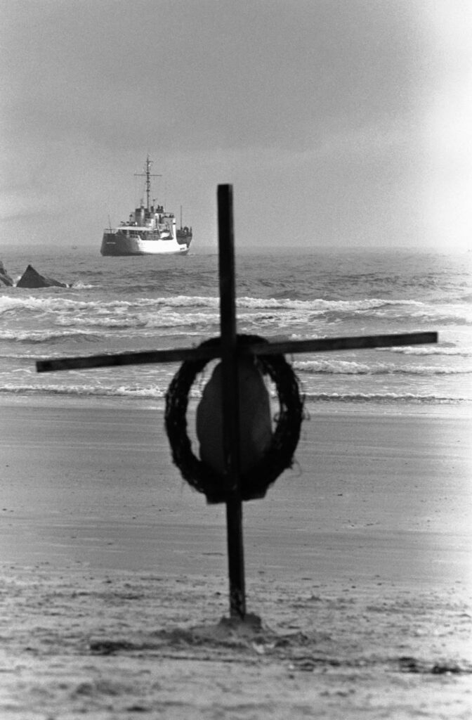 Uma cruz e uma coroa de flores estão na praia enquanto um navio da Guarda Costeira parte em busca de destroços da explosão. Fevereiro de 1986. (Imagem: Jim Neihouse / AP).