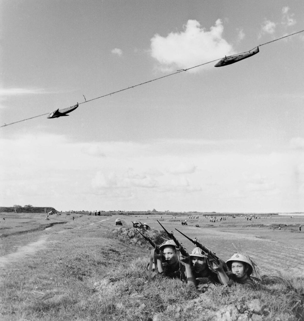 Utilizando alvos aéreos, uma companhia de milícia pratica tiro à frente de aeronaves em alta velocidade em Thanh Tri. Mesmo usando rifles antiquados da Segunda Guerra Mundial como estes, os vietnamitas conseguiram danificar ou derrubar muitas aeronaves americanas. Este grupo de milícia, a Companhia 6 da Comuna de Yen My, conquistou o título de "Excelente Milícia" por três anos consecutivos. 1965. (Foto de Minh Dao).